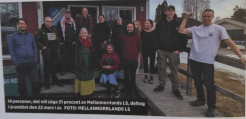 Gruppbild utanför ett rött hus. Fjorton personer, en sitter på trappan i röd tröja och blå klänning med väska i famnen.