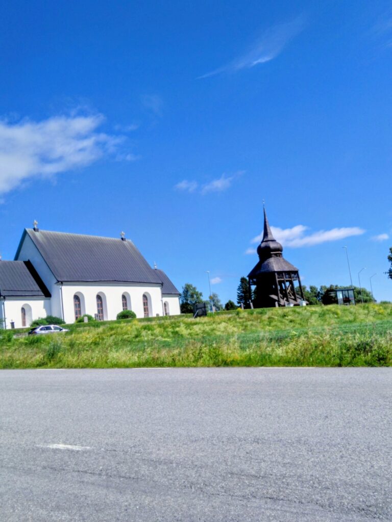Frösö kyrka i vitt med fristående klockstapel mot klarblå himmel, sett från busshållplatsen. Foto: Marlene Tyckosdotter